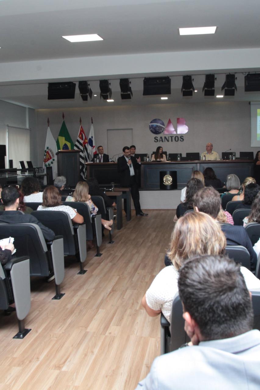 Auditório lotado de pessoas assistindo a uma palestra. Ao fundo, um homem fala ao público. #Pracegover