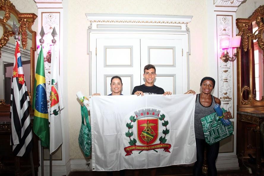 Os três atletas seguram bandeira da Cidade de Santos no salão nobre da prefeitura. #pracegover