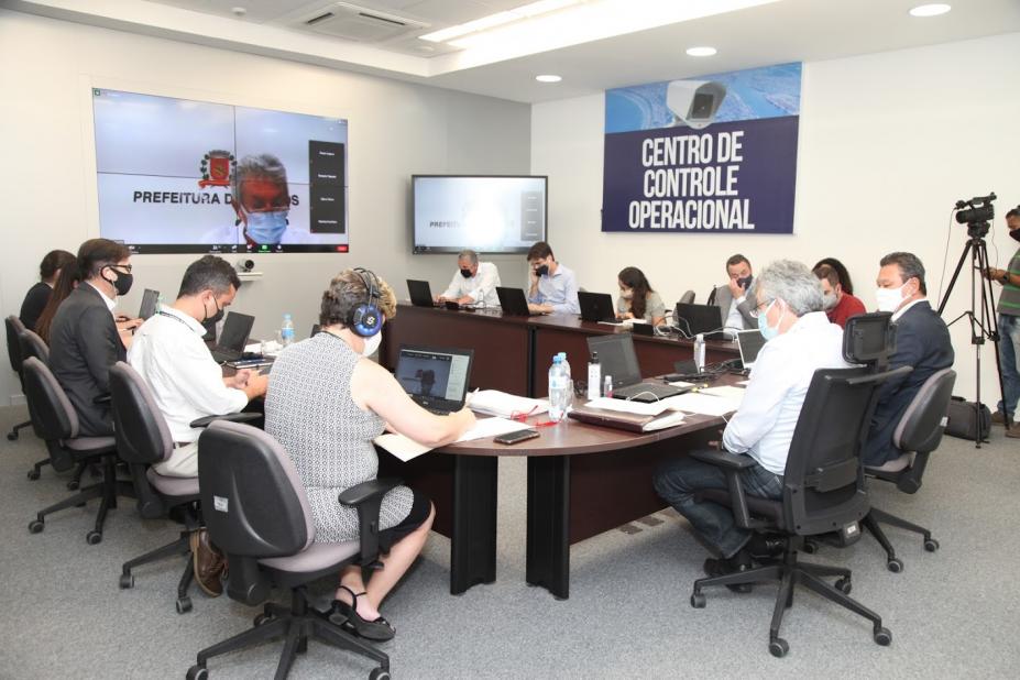 Várias pessoas sentadas em torno de uma mesa. Na parede está fixado um telão com a imagem de um dos integrantes da reunião. #Paratodosverem