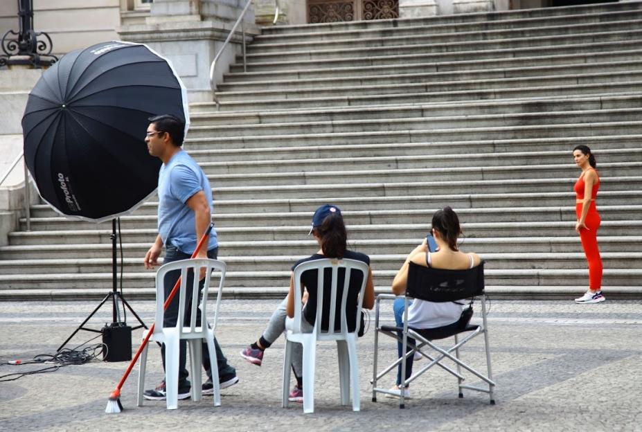 equipamentospara filmagem e fotografia dispostos em praça. Ao fundo, numa escadaria há uma mulher parada, de perfil, indicando que é a modelo alvo da filmagem. #paratodosverem