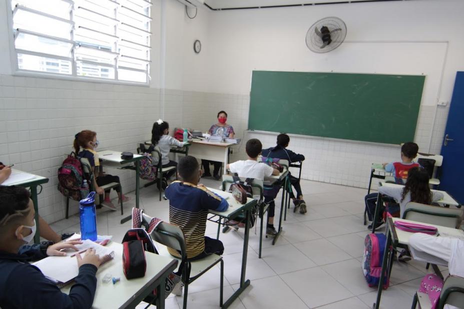 sala de aula com professor ao fundo e alunos sentados às carteiras. Lousa e ventilador afixados na parede ao fundo. #paratodosverem