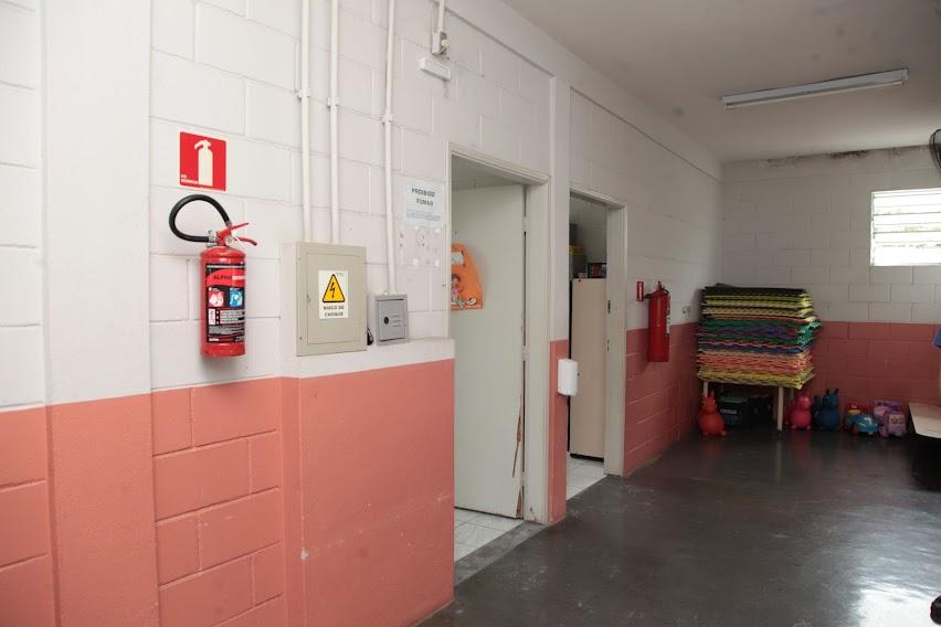 corredor de escola com extintor pendurado em parede à esquerda. Há duas portas na lateral esquerda da parede. Ao fundo, colchonetes empilhados. #paratodosverem