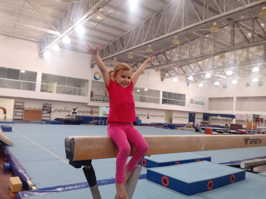 Criança com os braços estendidos em cima da trave de ginástica #paratodosverem