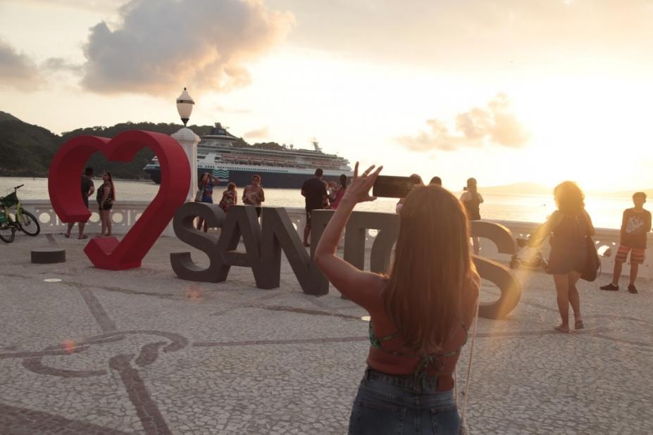 mulher etá diante de monumento Santos fotografando. Há um navio passando ao fundo. #paratodosverem