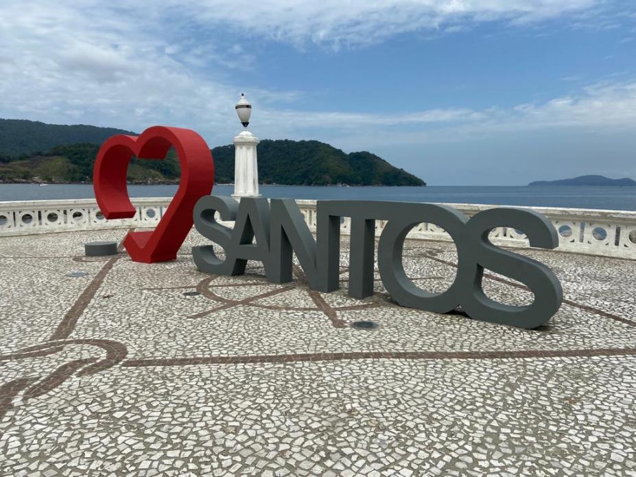 a escultura em um recuo na orla, com o coração em vermelho ao fundo e a palavra santos em cinza. #paratodosverem