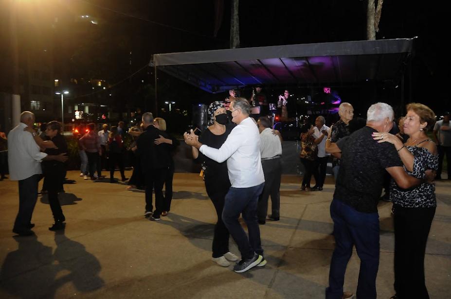 pessoas em duplas estão dançando em pista na orla. Ao fundo, um palco. Não é possível ver os  músicos. #paratodsoverem