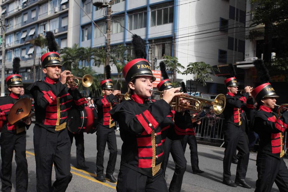 Banda desfila em rua. Músicos uniformizados tocam instrumentos de sopro. #Pracegover