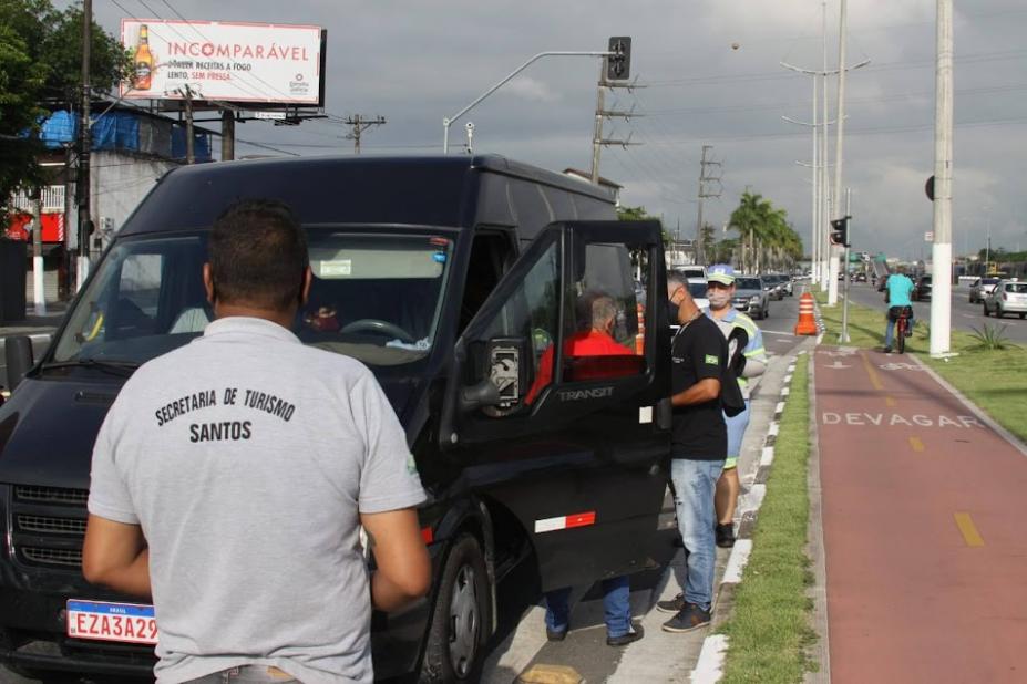 HOMEM COM CAMISETA COM ESTAMPA DO NOME DA SECRETARIA DE TURISMO ESTÁ DE COSTAS, DIANTE DE UMA VAN. A van está de porta abeerta, com duas pessoas ao lado conversando. Eles estão em uma avenida, ao lado de uma ciclovia. #paratodosverem