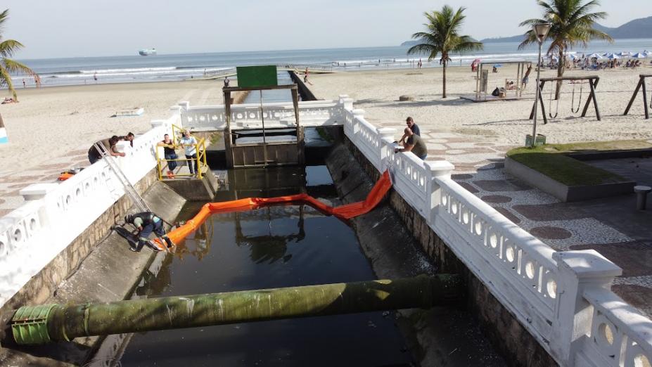 barreira é instalada dentro de canal junto à orla. Dois homens estão de cada lado do canal, sendo um dentro e outro fora. ambos estão segurando as pontas das barreiras. #paratodosverem