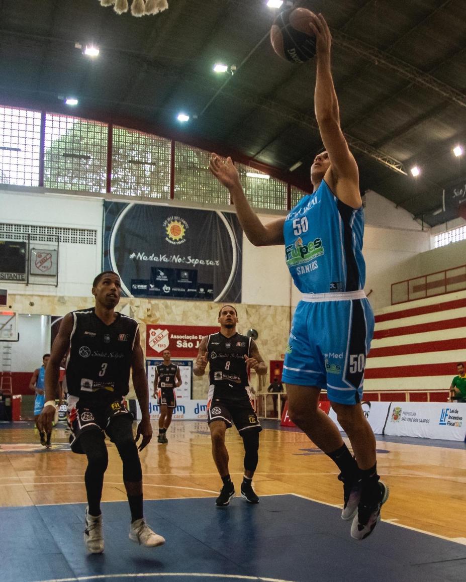 atleta da equipe de santos em primeiro plano está no ar, levando a bola ao cesto. Atrás, dois jogadores da equipe adversária