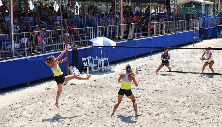 duas equipes jogam beach tennis, com duas duplas em lados opostos da rede ao centro da quadra de areia. São mulheres. Elas seguram raquetes e fazem movimentos do jovo. Ao lado esquerdo, a arquibancada. #paratodosverem