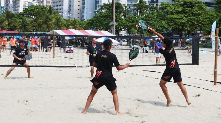 duas duplas jogam beach tennis em quadra na areia da praia. Dois atletas estão de costas segurando e movimentando as raquetes. Outros dois, do outro lado da rede, também seguram as raquetes. #paratodosvere