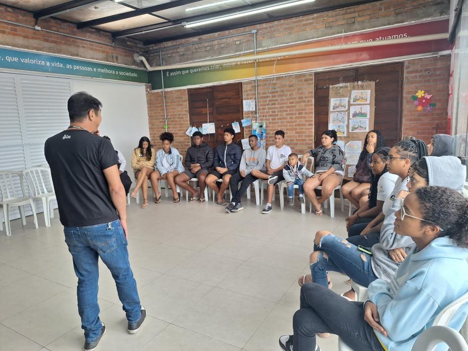 homem está em pé, de costas para a foto, falando a grupo de jovens que estão sentados em um auditório. #paratodosverem 
