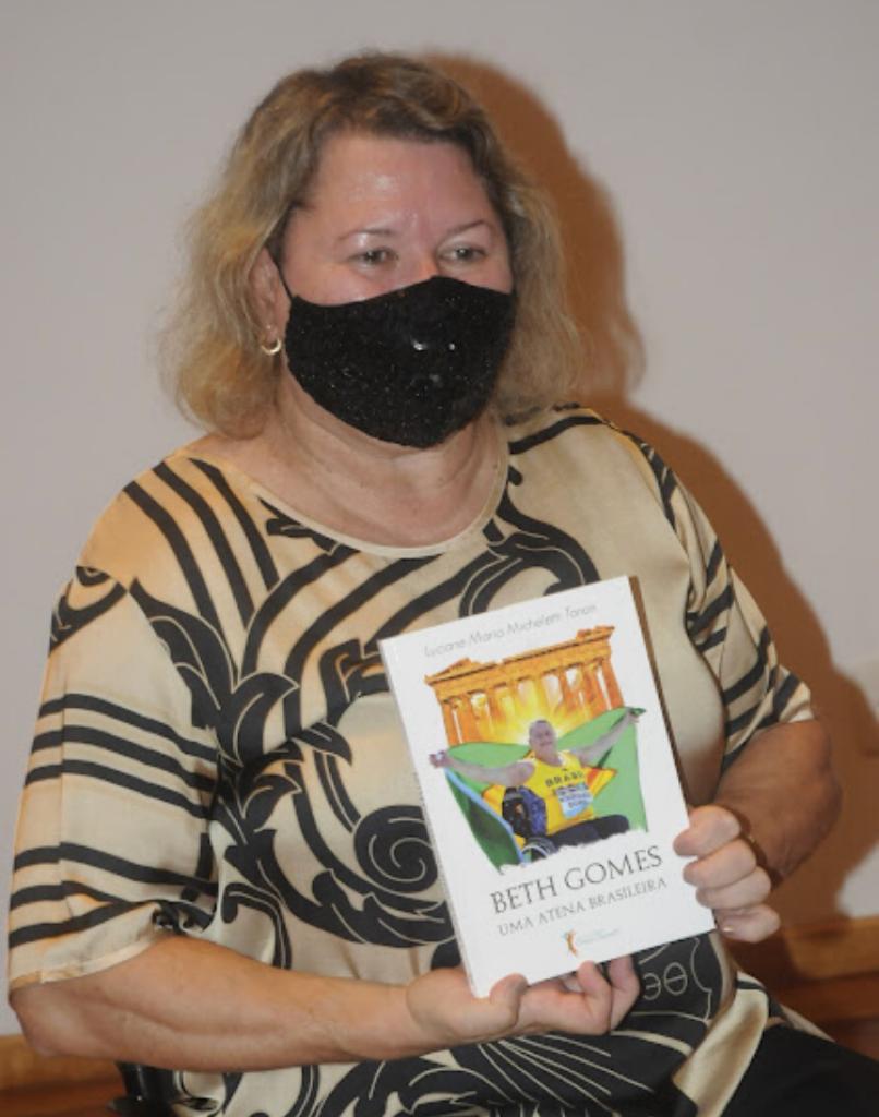 Betha Gomes está sentada e usando máscara. Ela segura o livro com as duas mãos. #paratodosverem