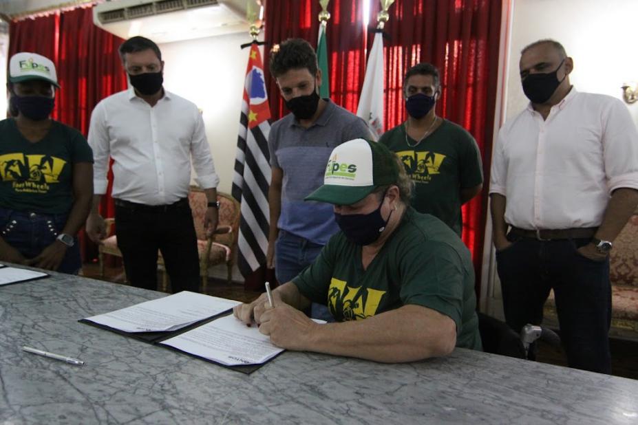 atleta assina documento e demais autoridades ao fundo observam #paratodosverem