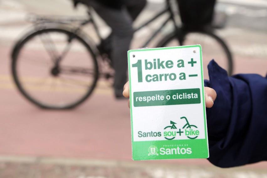 close de mão segurando pequena placa onde se lê uma bike a mais um carro a menos. Ao fundo, uma bicicleta passando. #paratodosverem