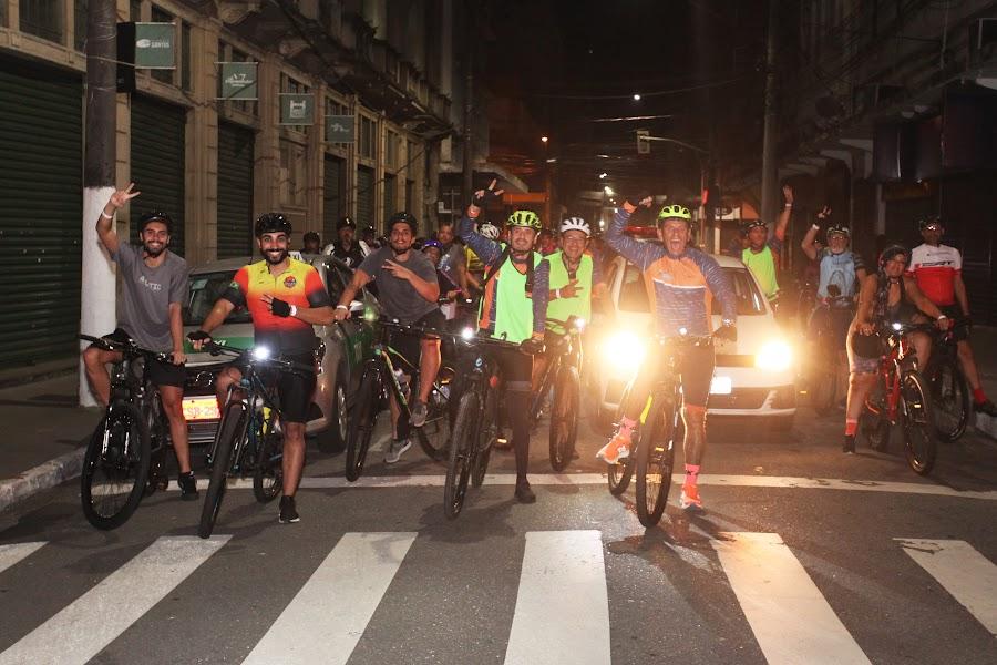 ciclistas param diante de faixa de pedestre para foto. É noite. Eles acenam. #paratodosverem
