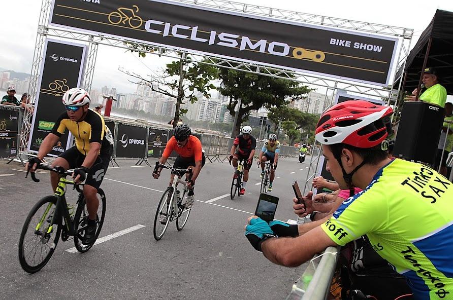 Ciclistas pedalam na avenida enquanto ciclista tira foto pelo celular do lado de fora da prova. #pratodosverem