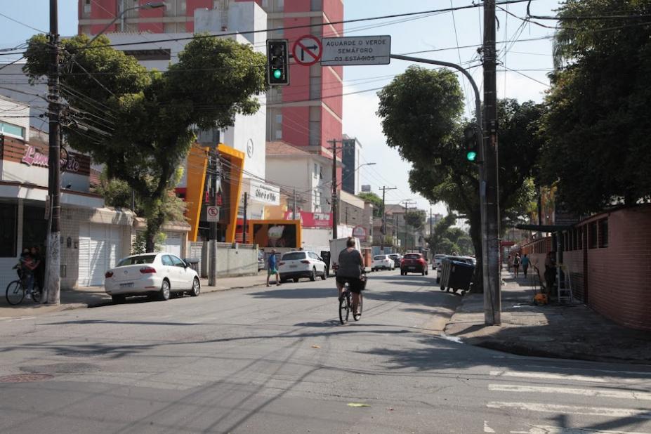 trecho de rua com calçadas, carros estacionados dos dois lados e uma mulher de bicicleta indo adiante da foto. #paratodosverem