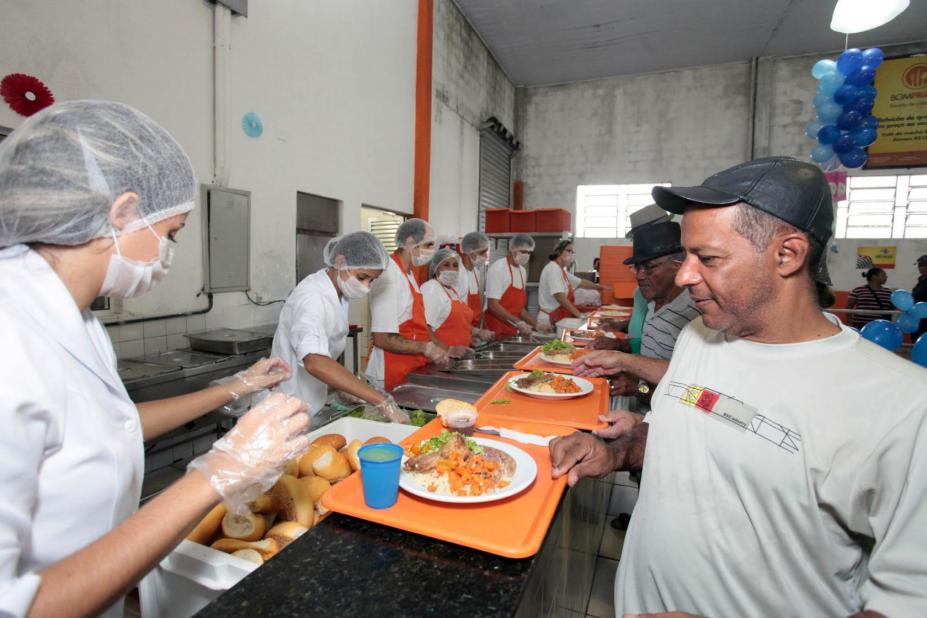 Imagem de uma unidade do Bom Prato, com pessoas atrás do balcão servindo os usuários do serviço. #paratodosverem