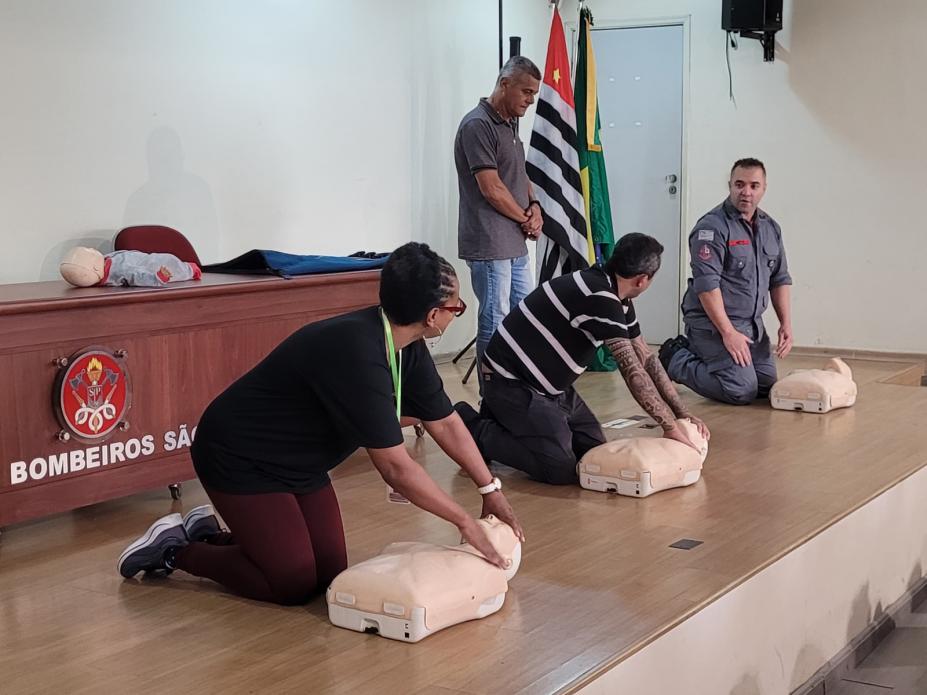 três pessoas estão ajoelhadas no chão massageando peito de boneco que simula uma pessoa. Uma dessas pessoas é o bombeiro que orienta o procedimento. #paratodosverem 