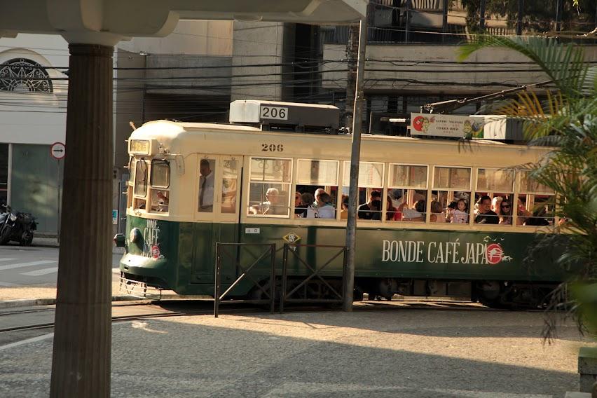 bonde fazendo curva em esquina do centro de santos. Na lateral do veículo está escrito Bonde Café Japão. #paratodosverem