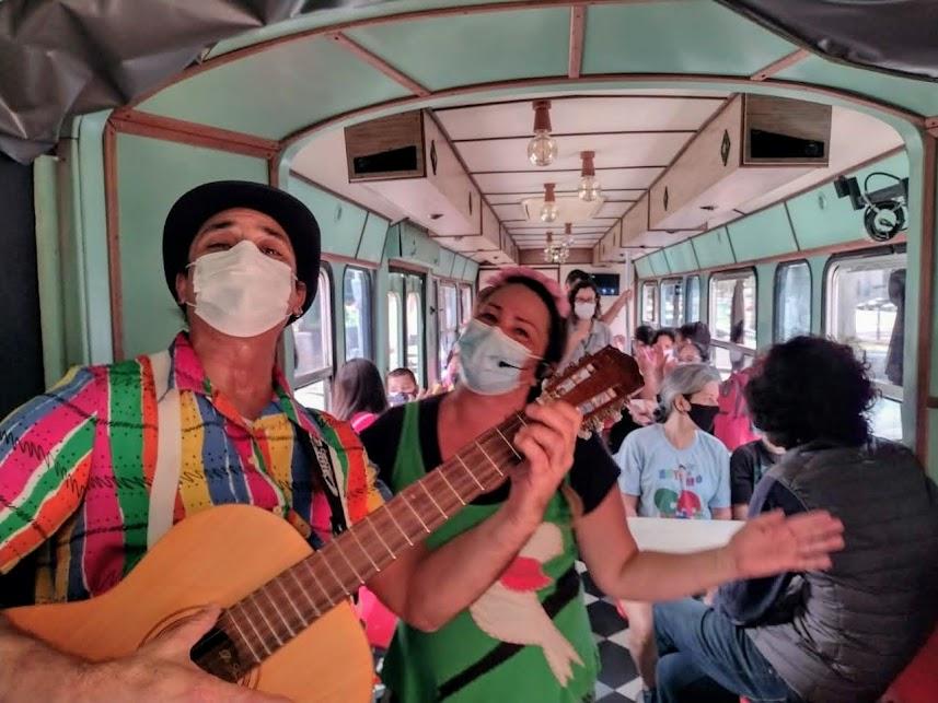 homem tocando violão ao lado de mulher. Eles estão em primeiro plano dentro do bonde. Ao fundo se vê o público. #paratodosverem