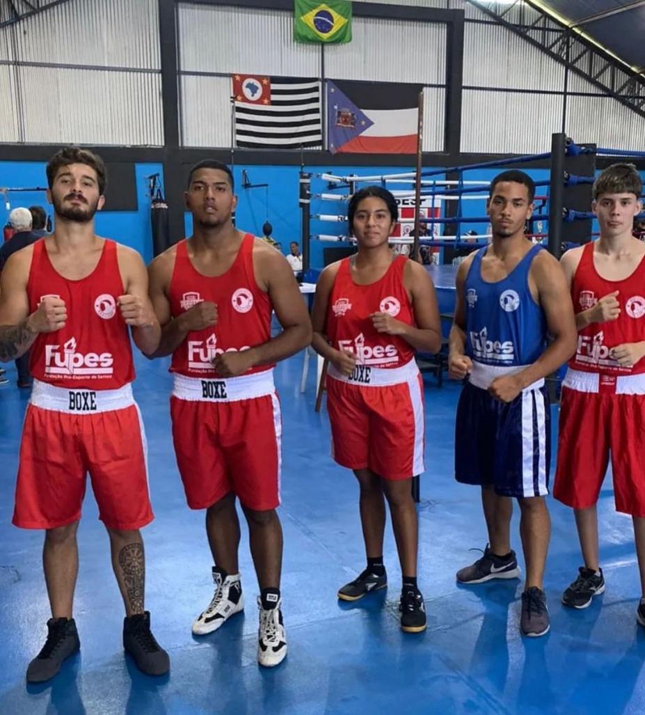 cinco boxeadores, sendo quatro homens e uma mulher ao centro, posam para a foto com as mãos em gesto de luta. #paratodosverem