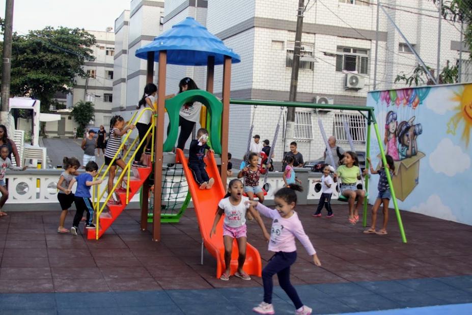 crianças estão subindo na escada e descendo o escorregador. Elas se divertem. Ao fundo, outras crianças estão brincando nos balancês. #paratodosverem