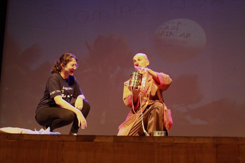 Mulher e palhaço estão lado a lado em palco. eles estão agachados. A mulher ri. #paratodosverem