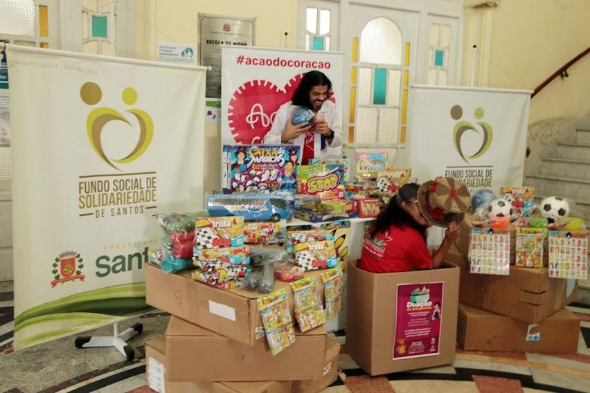 vários brinquedos dentro de caixas e sobre mesas. Um homem está atrás da mesa e à frente de banners. Ele ri. Á frente, uma mulher está dentro de uma caixa e cobre parte do rosto com um chapéu.#paratodosverem