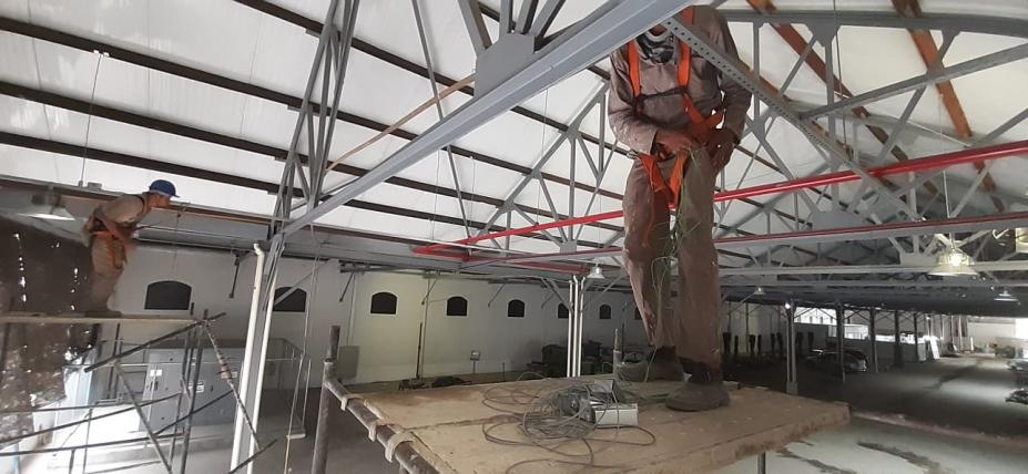 em primeiro plano, homem está sobre andaime trabalhando na estrutura interna do telhado. #paratodosverem