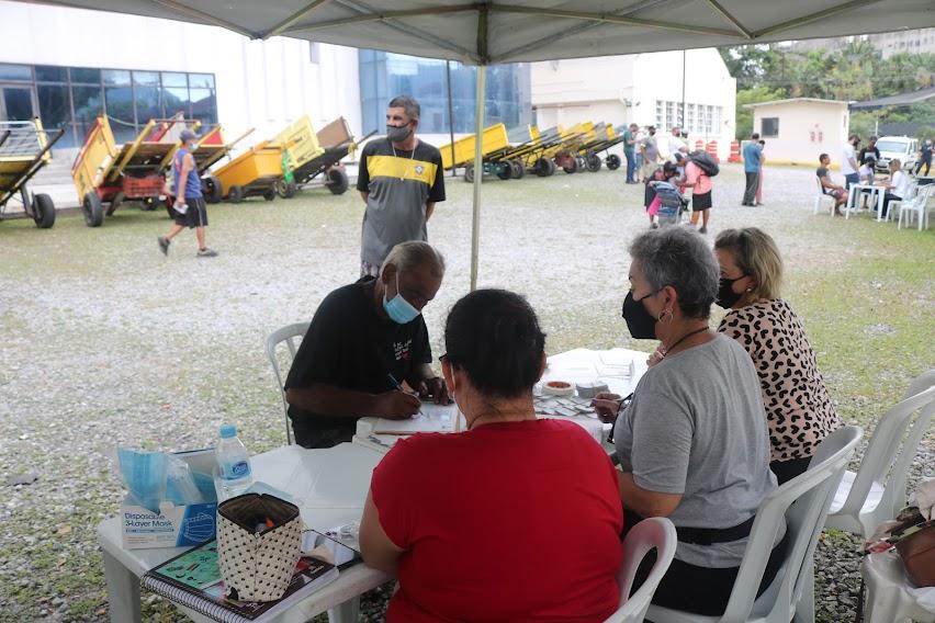 homem está sentado diante de mesa, sendo atendido por três pessoas. Ao fundo, vários carrinhos de coletores. #paratodosverem