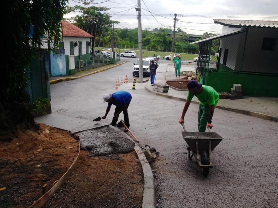 homens passam concreto em trecho de calçada. Um deles aplica a massa. O outro está segurando um carrinho de mão. #paratodosverem 