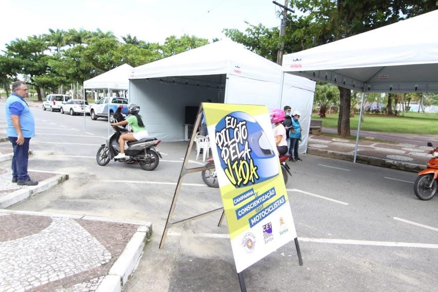 placa na rua onde se lê Eu piloto pela vida. Ao fundo, três tendas estão montadas. Há um agente da CET e uma motocicleta sendo conduzida por uma mulher. #pratodosverem