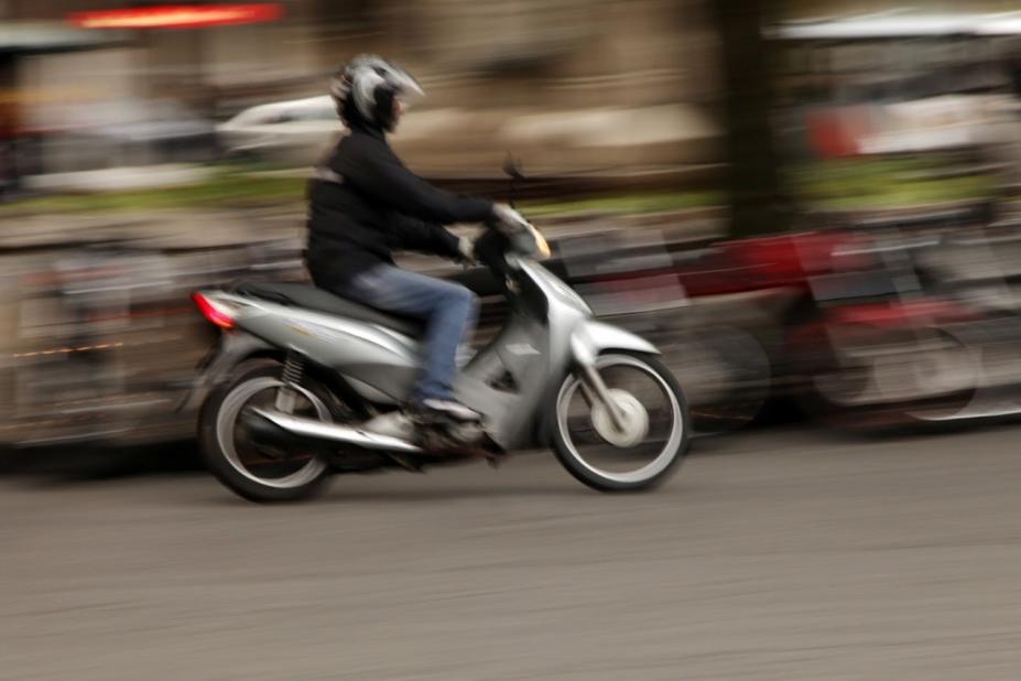 Imagem de homem em moto prateada passando por via pública com imagem de fundo desfocada. #pratodosverem
