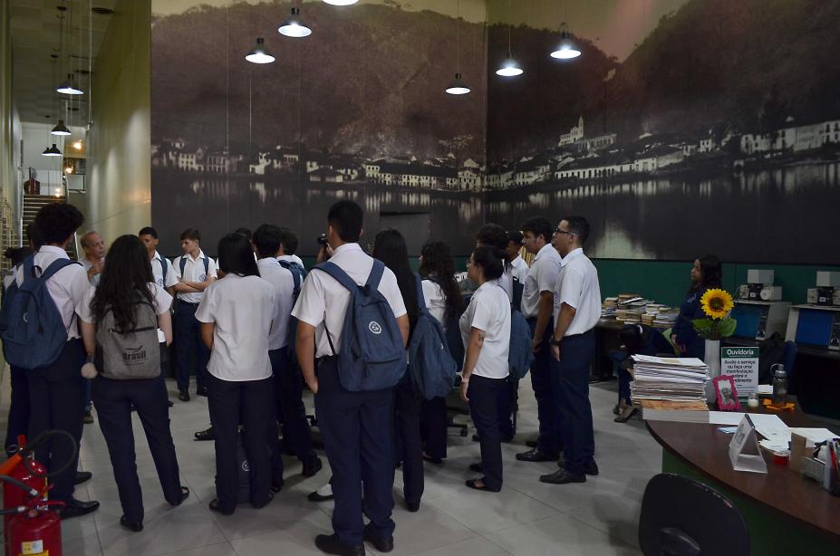 jovens estão em pé em sala com grande foto de Santos antiga ao fundo. #paratodosverem