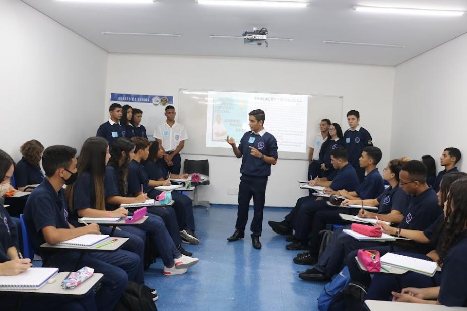 aprendizes estão sentados em sala. Alguns estão em pé. Um deles, o principal, no meio da sala, fala ao grupo. #paratodosverem