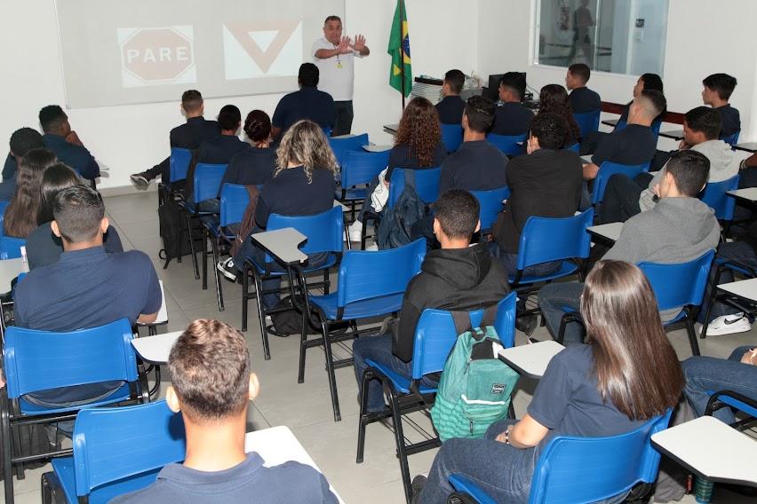 sala está cheia de jovens sentados assistindo palestra. Homem fala ao fundo. No telão que está atrás dele se vê dois sinais de trânsito. #paratodosverem