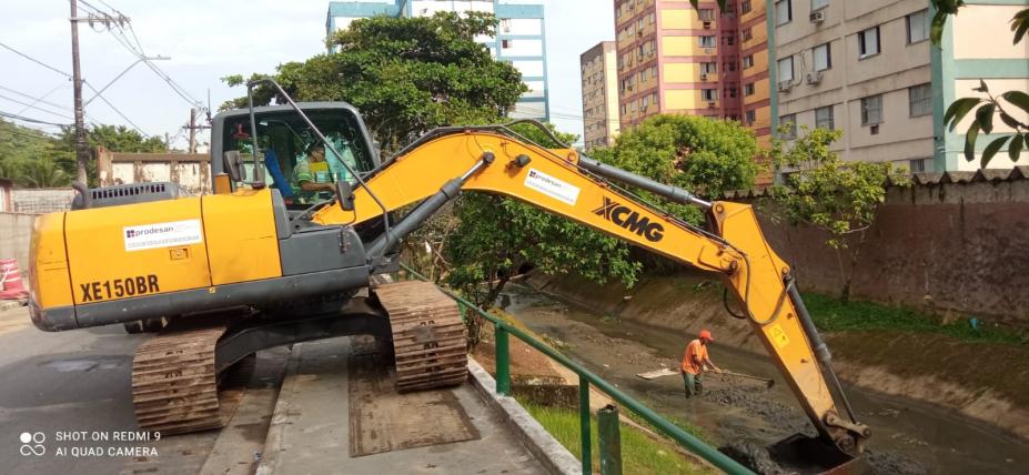 escavadeira está na ocupando parte da rua e calçada e com o braço pra dentro do canal removendo detritos. Ao fundo, no canal, há um homem também coletando detritos. #paratodosverem