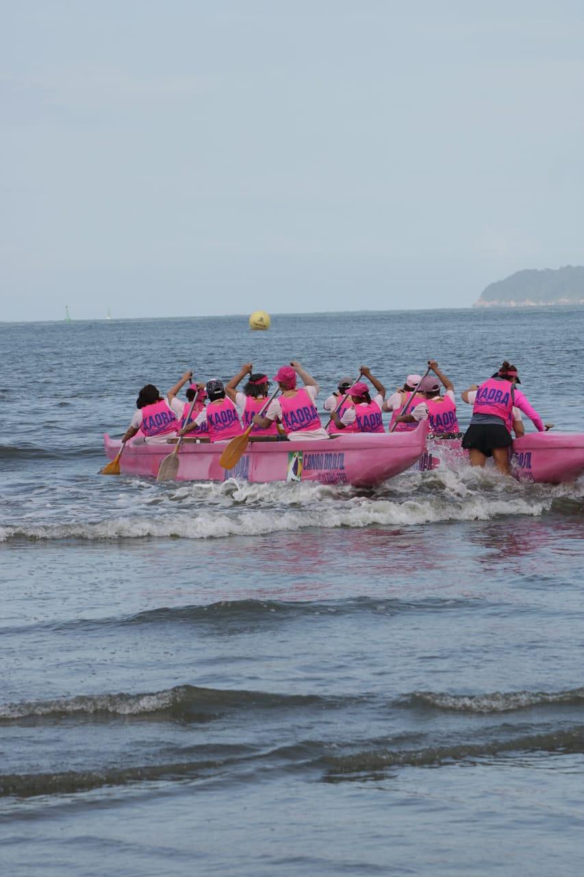 Canoa havaiana na cor rosa ocupada com mulheres vestidas de rosa ingressa no mar. #pracegover