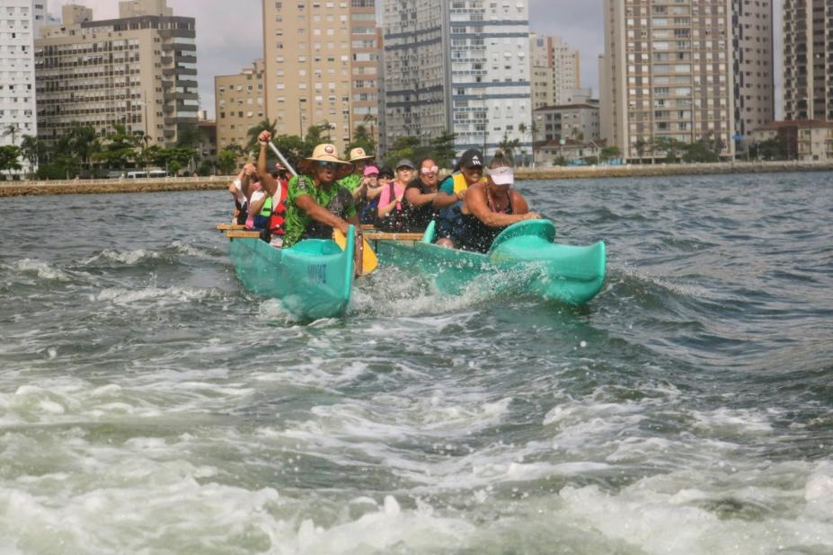 mulheres remam na água dentro da canoa. Há movimentação de ondas. #paratodosverem