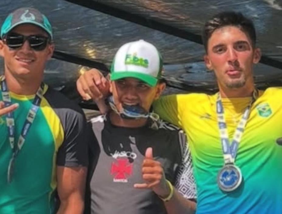 trés atletas posam para foto abraçados. O do meio segura uma medalha