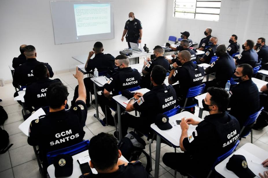 Guardas municipais uniformizados estão sentados em cadeira de sala de aula atentos a um homem que fala a todos, no fundo da classe. Na parede está projetada a imagem de um texto. #paratodosverem