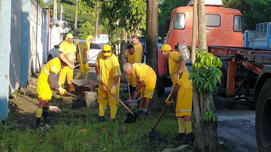 grupo de homens uniformizados capina calçada cheia de mato. Eles usam enxadas. #paratodosverem