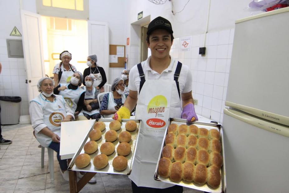homem segura duas bandejás com pães de cará recém-saídos do forno. Atrás dele as alunas estão sentadas. #paratodosverem