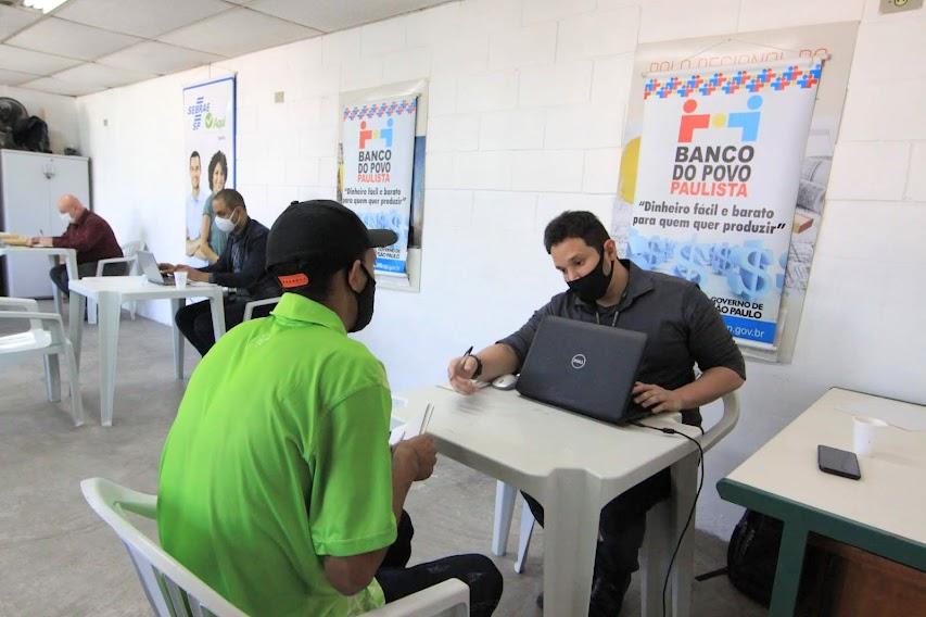 homem está sentado com laptop aberto à sua frente. Ele atende outro homem que está de costas para a foto. Ao fundo, um banner do banco do povo. #paratodosverem