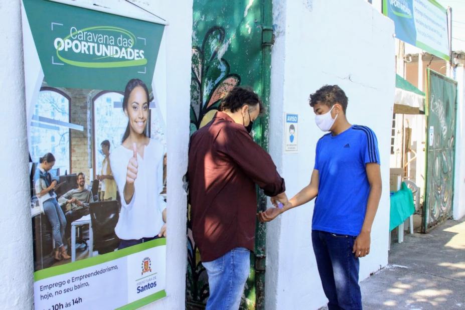 Homem mede a temperatura de jovem que vai entrar em sala. Na parede externa está pendurado um banner onde se lê Caravana das Oportunidades. #paratodosverem