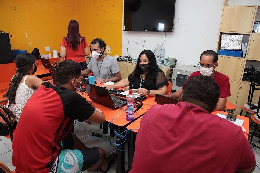 pessoas estão sentadas diante de mesa sendo atendidas por outras que estão diante de notebooks. #paratodosverem