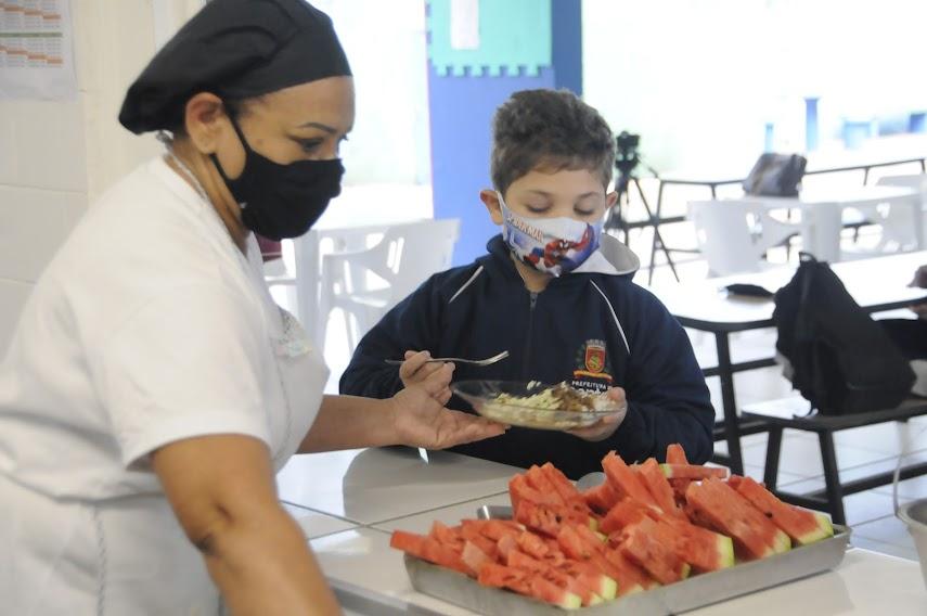 Merendeira coloca alimento para aluno, com pedaços de melancia em bandeja à frente. #pracegover